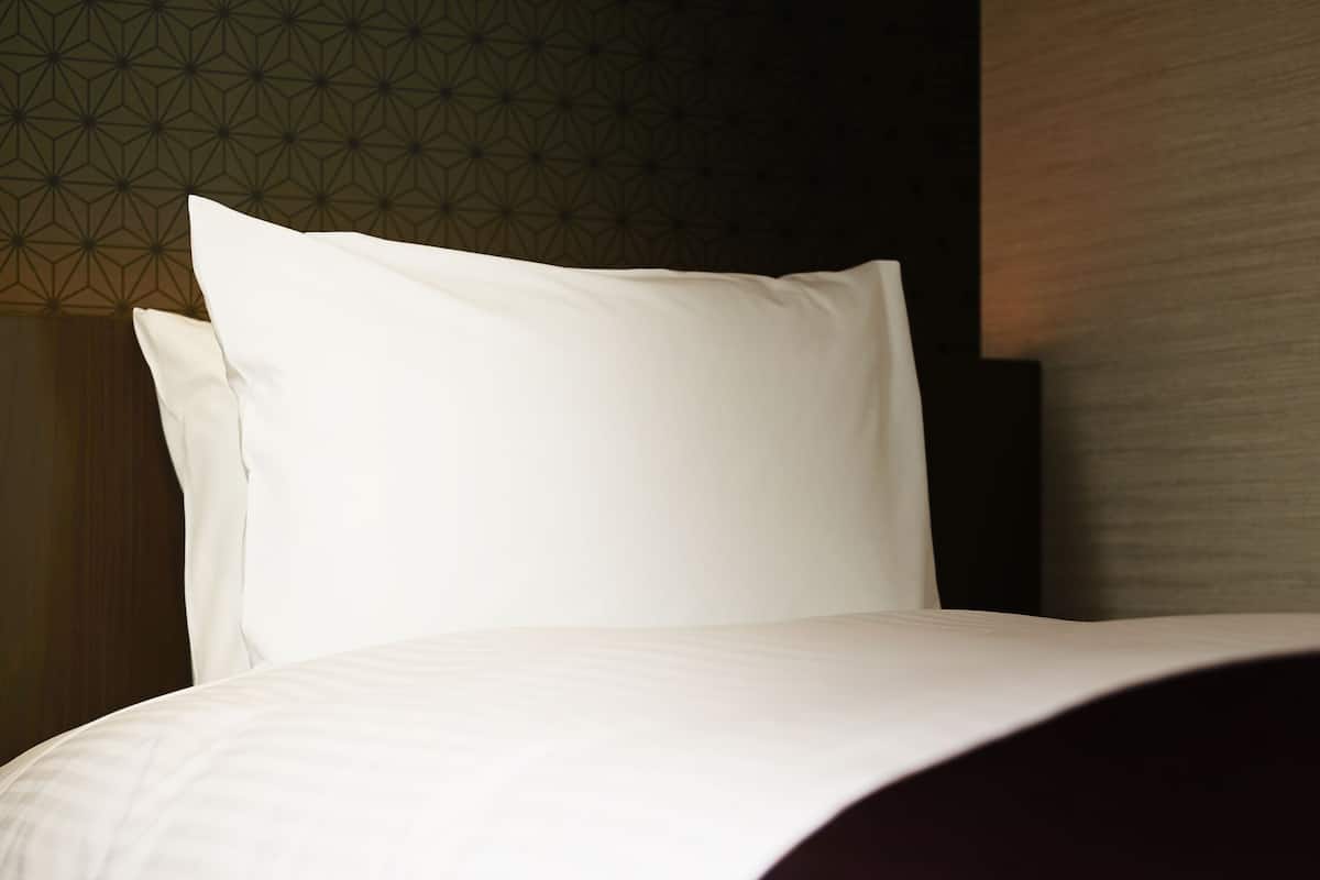 Una cama bien hecha con sábanas blancas y una manta oscura, frente a una pared con diseño geométrico.