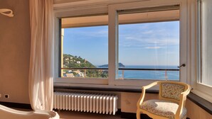 Una stanza con una grande finestra vista sulla costa, un termosifone sotto la finestra e una poltrona con motivo floreale.