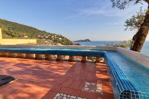 A swimming pool with a tiled edge, surrounded by terracotta tiles and a view of the sea and a hillside.