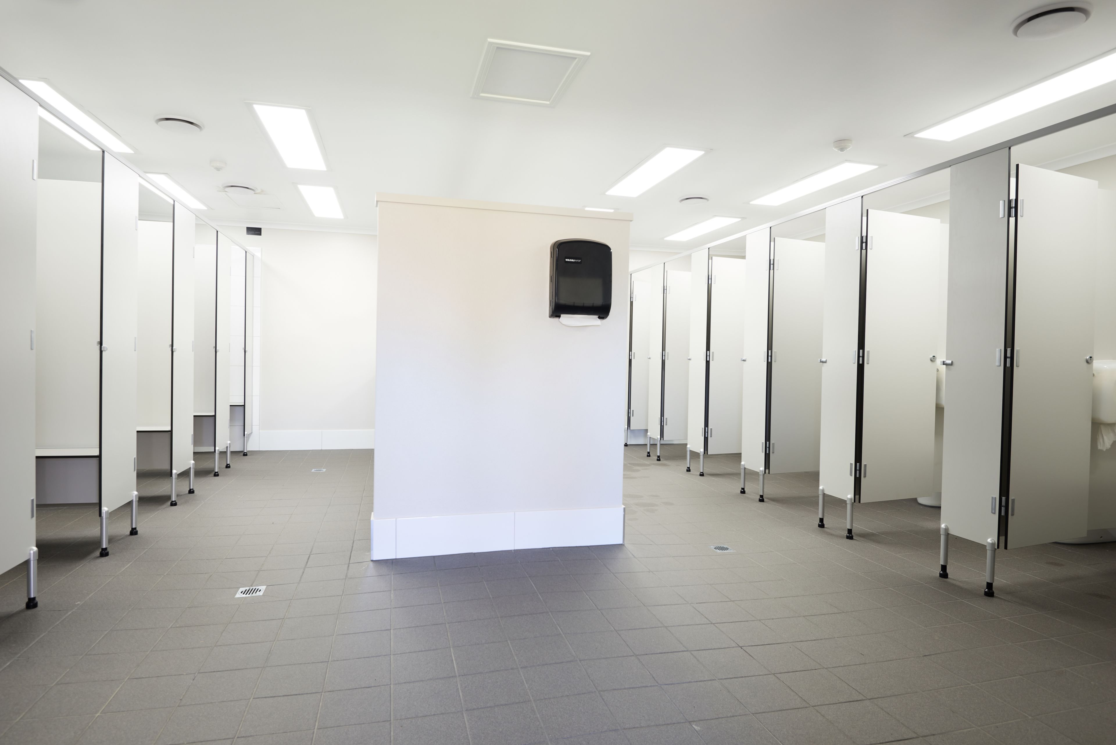 Baño público con cabinas múltiples, un secador de manos y piso de baldosas.