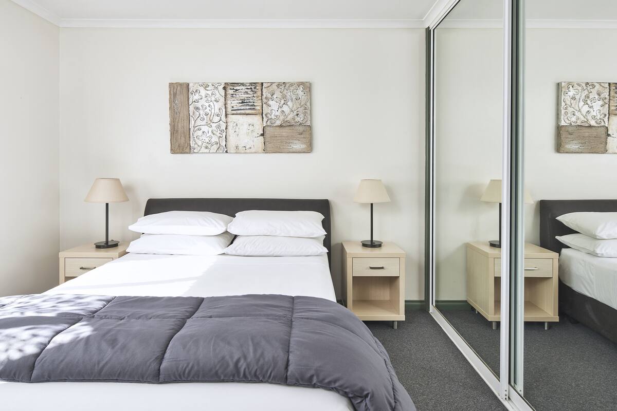 A neatly arranged bedroom with a bed, two bedside tables, a mirror, and wall art.