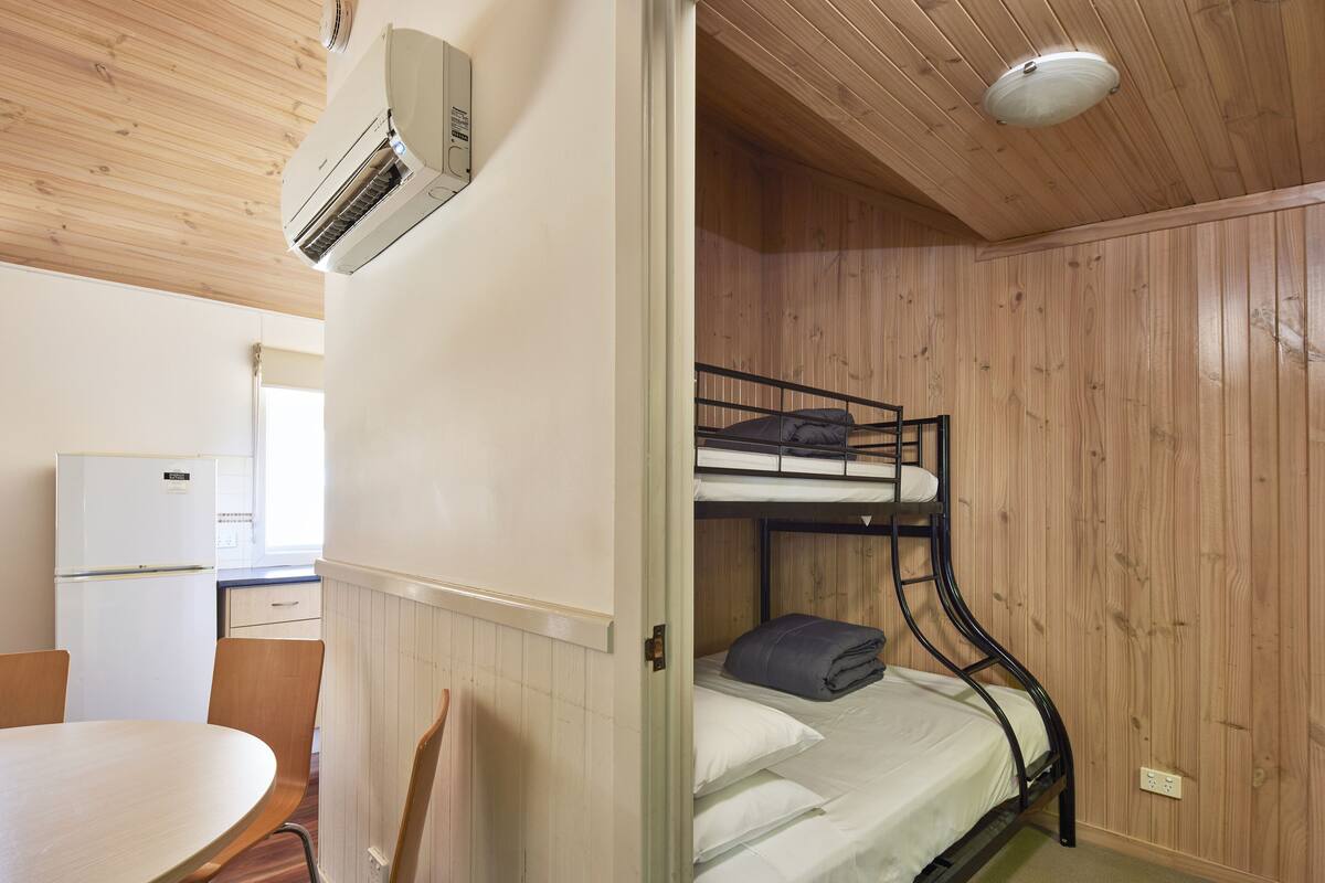 A bunk bed with a mattress and pillows, a white refrigerator, a wooden table, and a chair in a room with wooden walls and ceiling.