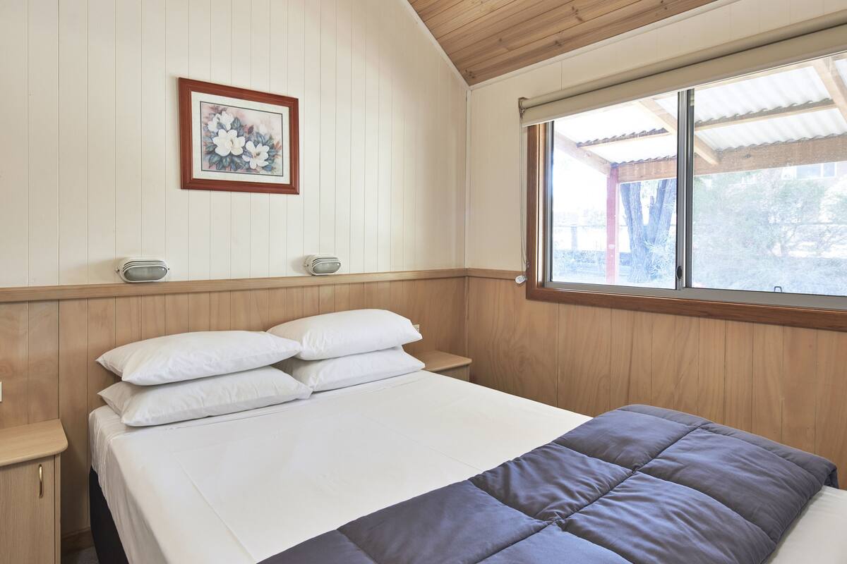 A bedroom with a bed, white walls, wooden paneling, a window, and a framed picture on the wall.