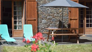 Un bâtiment en pierre avec des volets en bois, une table de pique-nique avec un parasol et deux fauteuils de jardin turquoise.