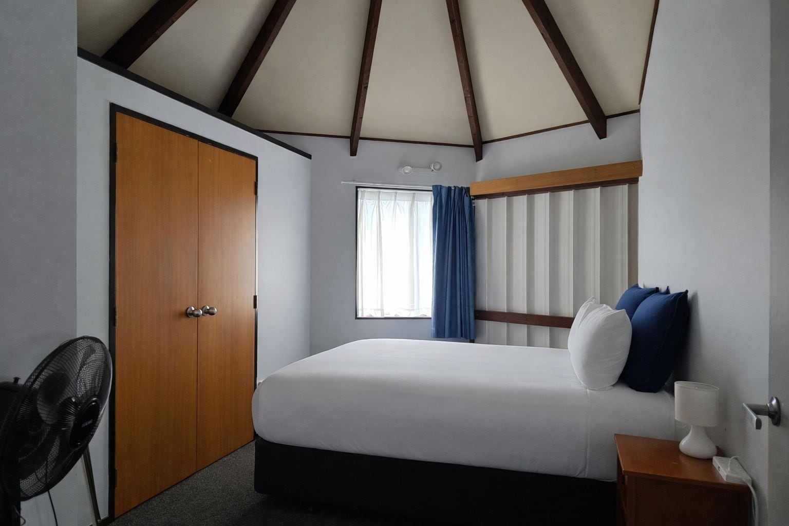 Una habitación de hotel con una cama, una puerta de madera, una ventana con cortinas y una mesita de noche con una lámpara.