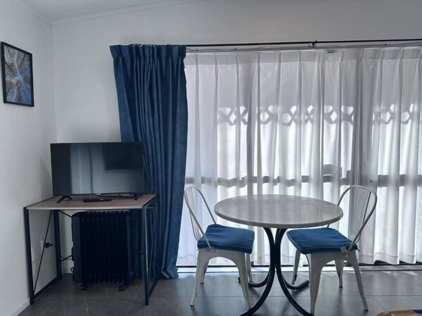 A room with a TV on a wooden stand, a round table with two chairs, and a large window with white curtains.