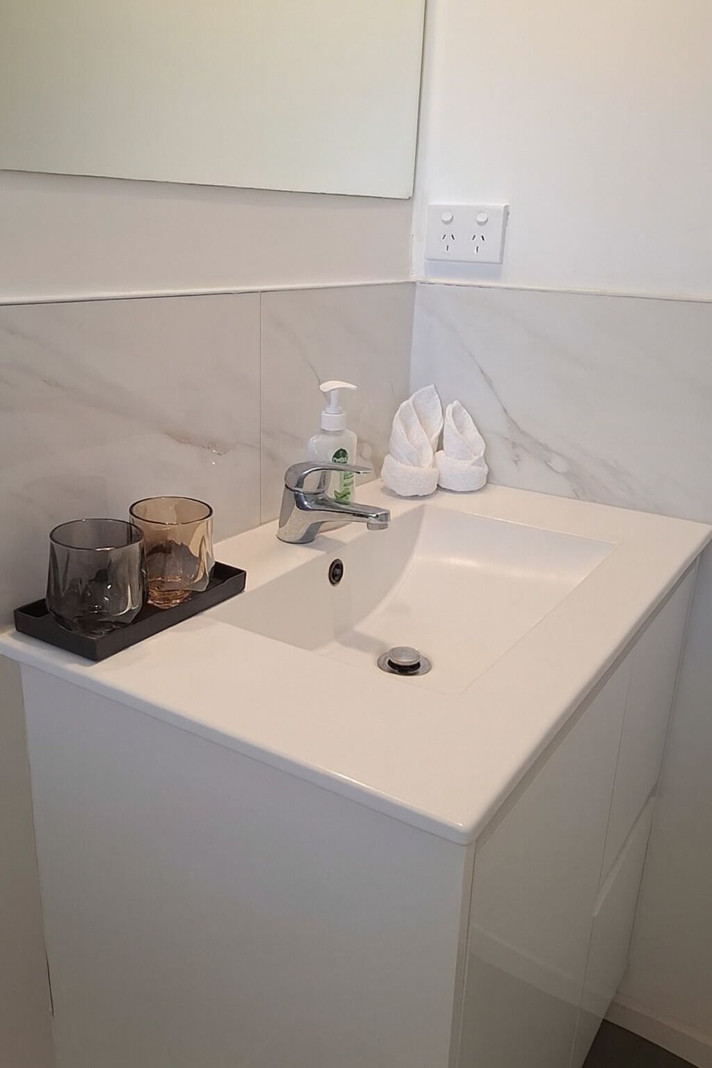 A bathroom with a white sink, a mirror, a soap dispenser, and a pair of white slippers.