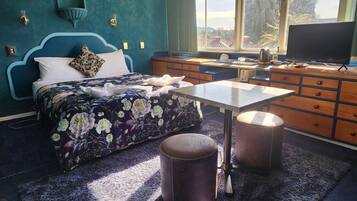 Ein Schlafzimmer mit einem Bett, einem Schminktisch, einem Tisch und einem Fenster mit Blick auf Bäume und Gebäude.