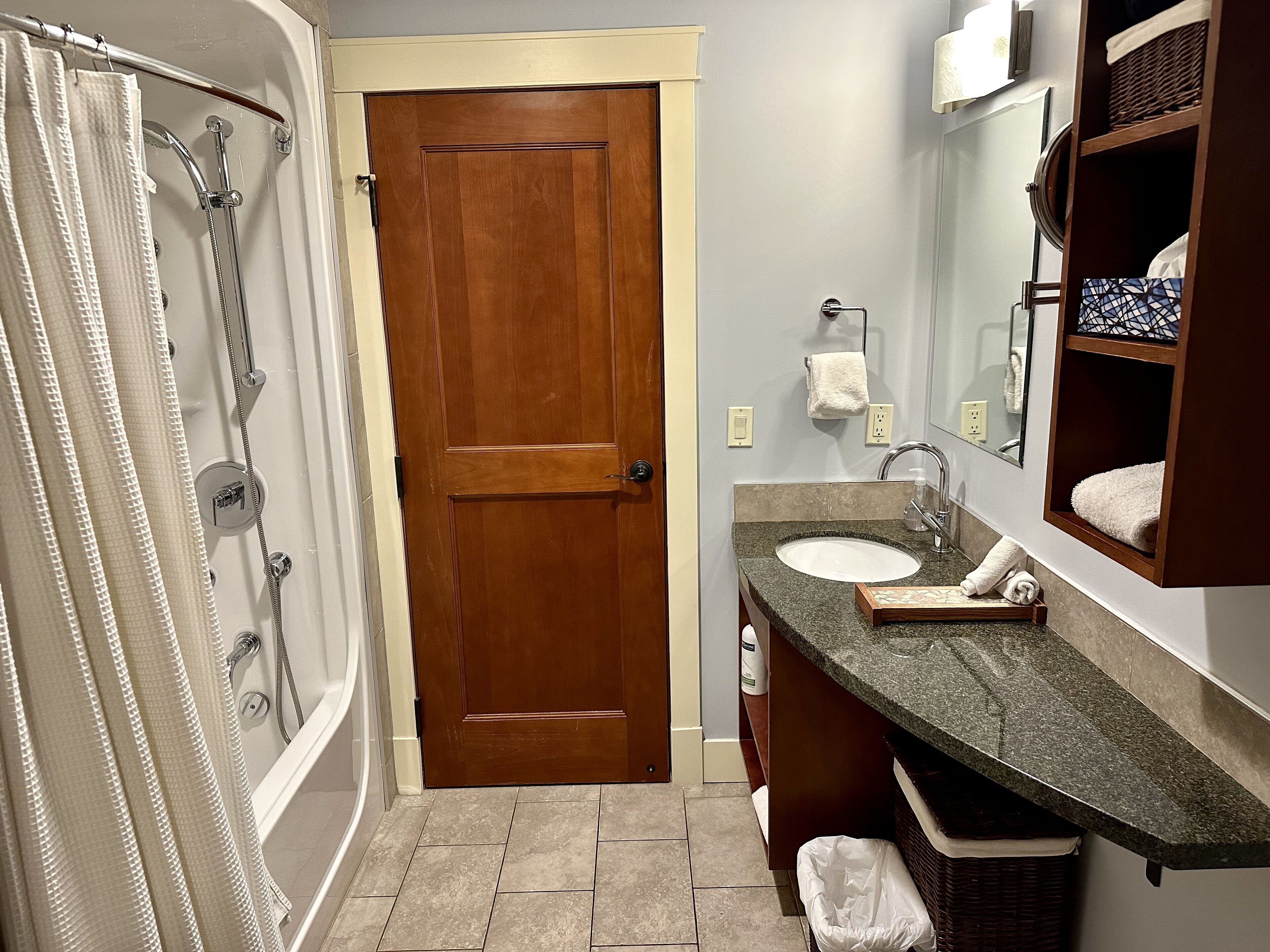 A bathroom with a wooden door, a shower with a curtain, a sink with a mirror, and a towel rack.