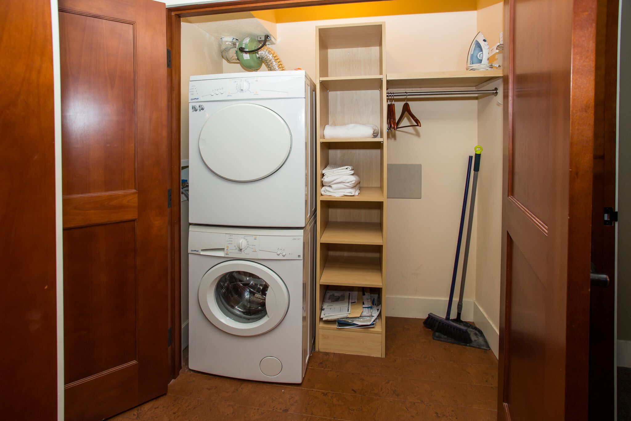 A laundry room with stacked washers and dryers, a broom, and shelves with folded towels.