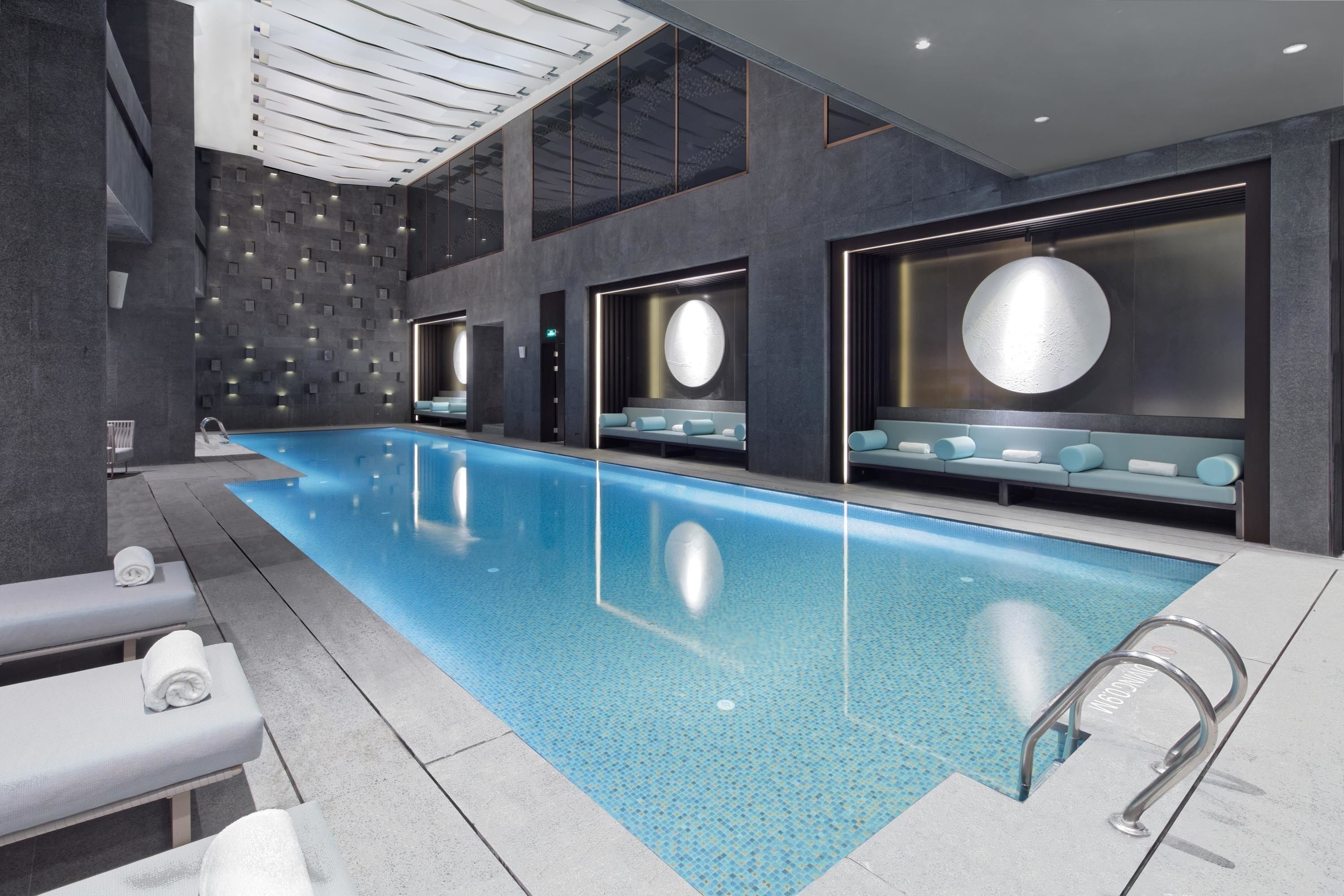 Una piscina interior moderna con superficie de agua cristalina, rodeada de zonas de descanso con asientos y toallas.
