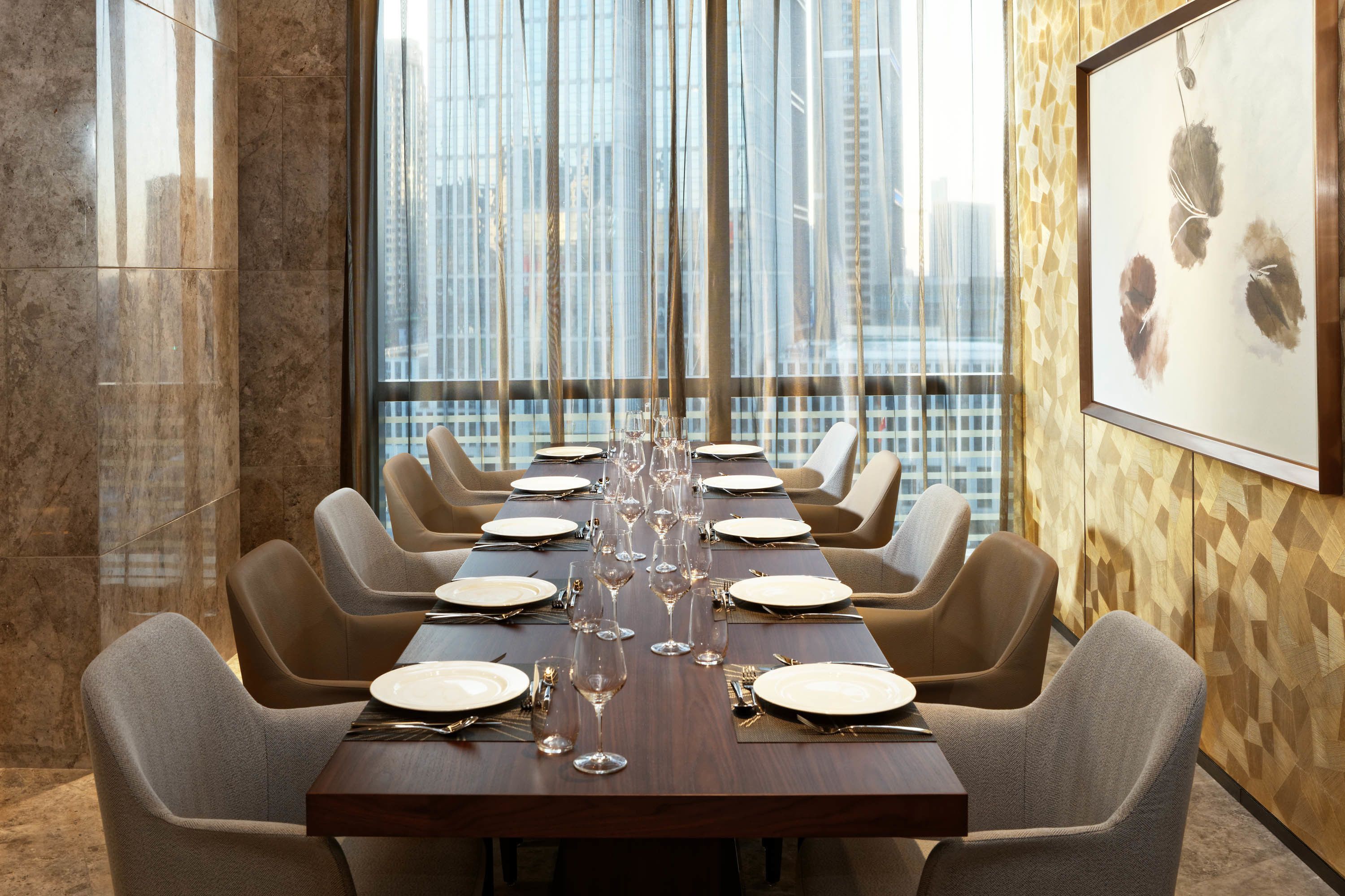 A dining area with a long table set for a meal, large windows with a city view, and a framed artwork on the wall.