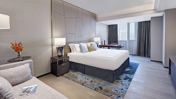 A modern hotel room with a large bed, a sitting area with a sofa, a small table with a lamp, and a view of the city through large windows.