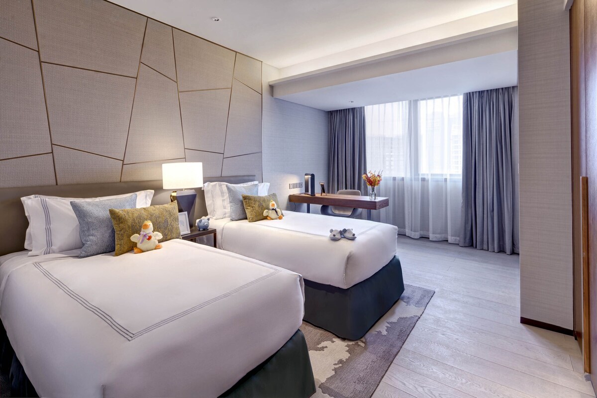 A hotel room with two beds, a nightstand, a desk, and a large window with curtains.