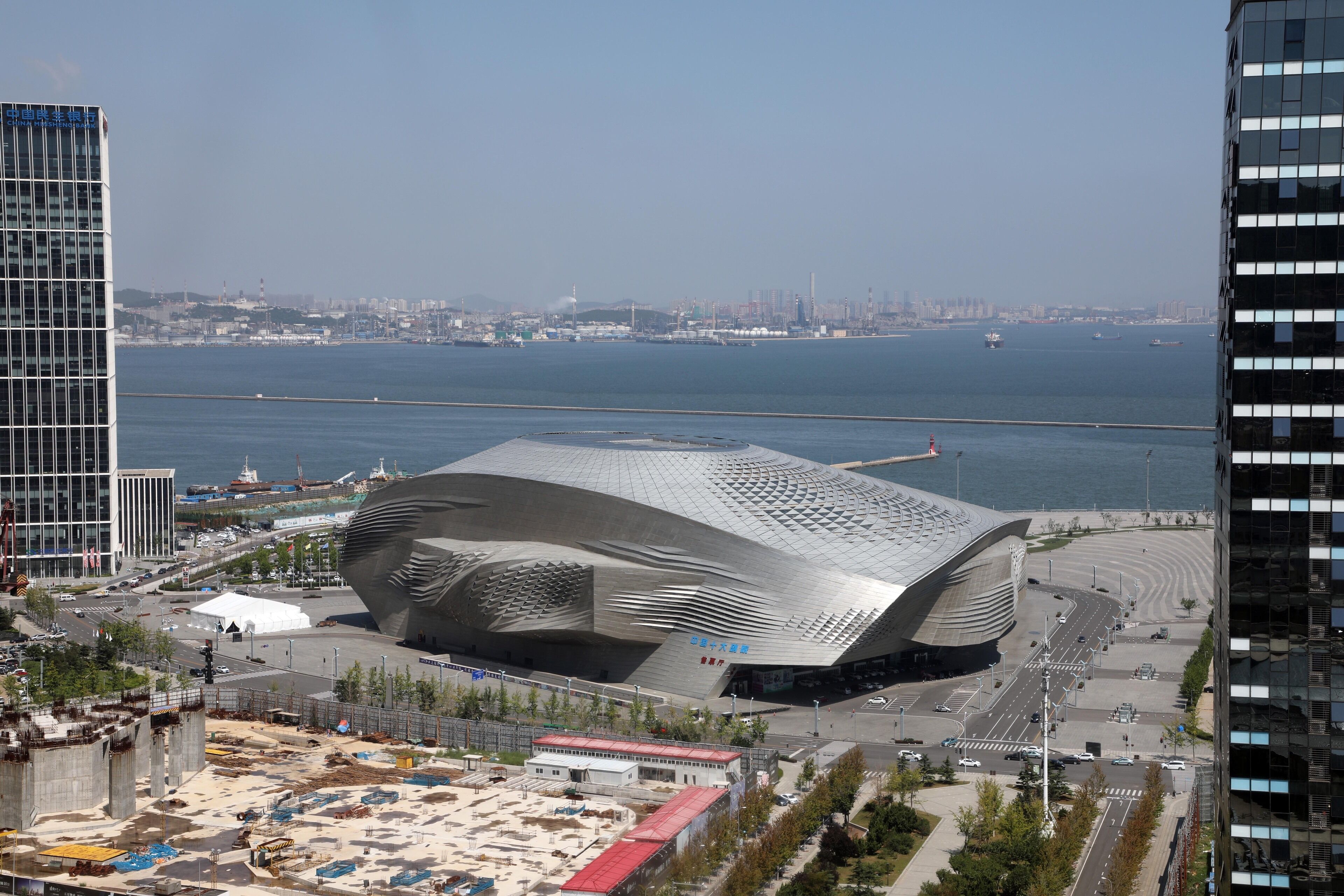 A modern architectural building with a unique curved design, situated near a waterfront with a view of the sea and other buildings in the background.