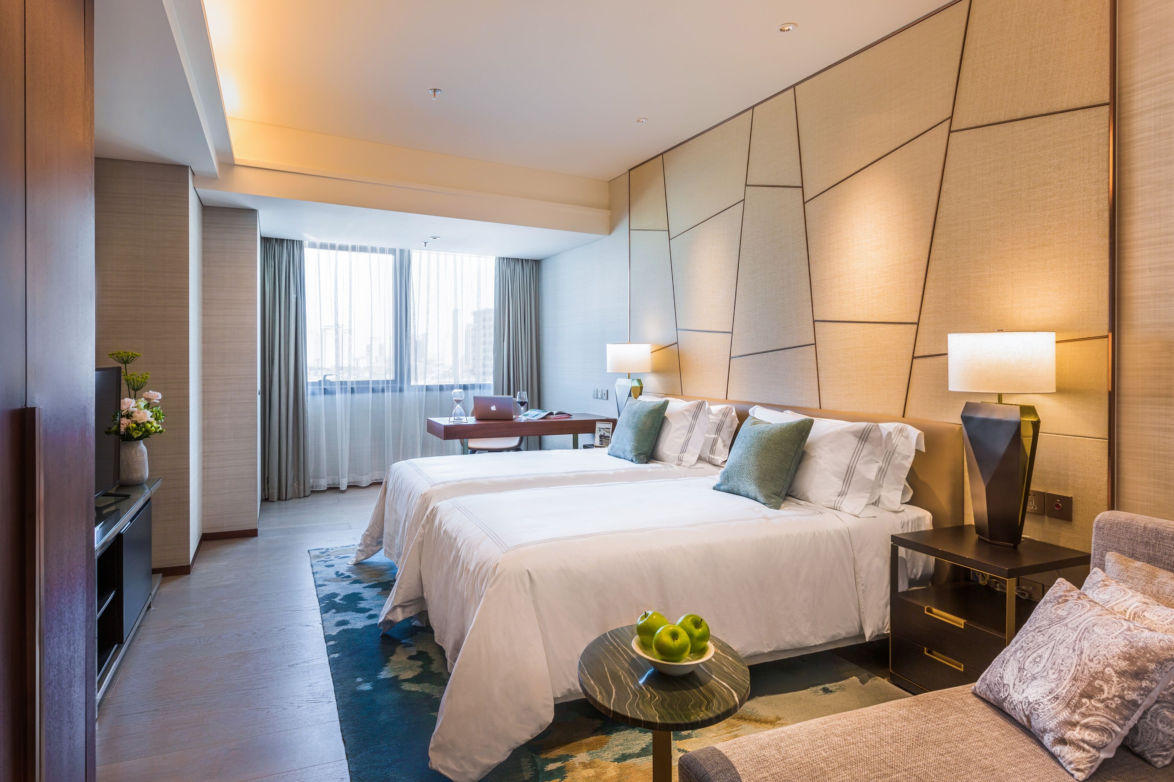 Une chambre d’hôtel avec deux lits, un bureau, une chaise, une lampe et une petite table sur laquelle se trouvent des pommes.