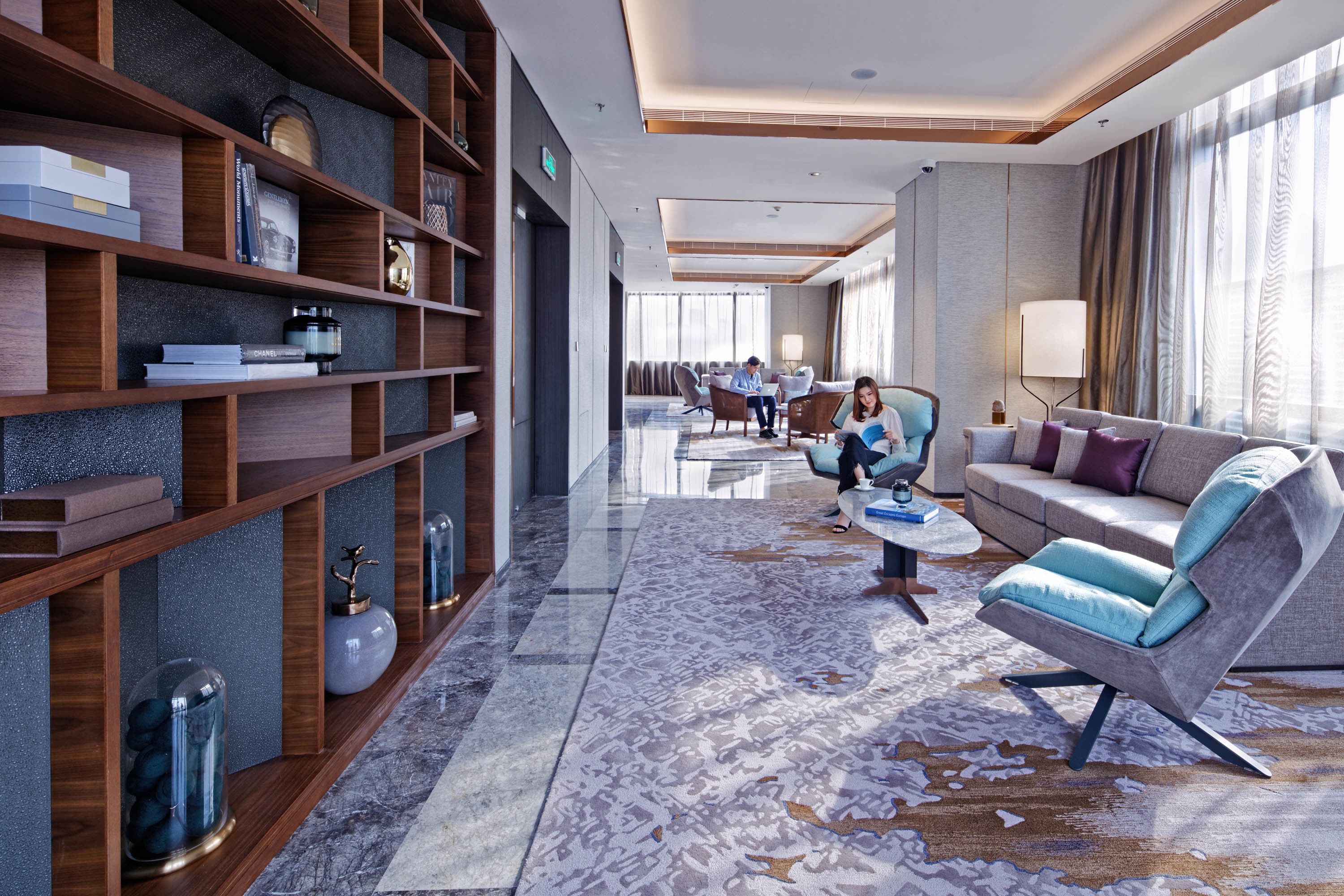 A modern hotel lobby with a spacious seating area, a large bookshelf, and a patterned carpet.