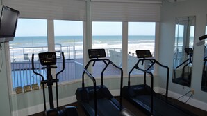 Un gimnasio con cintas de correr y equipo de ejercicio, amplias ventanas con vista a la playa y al océano.