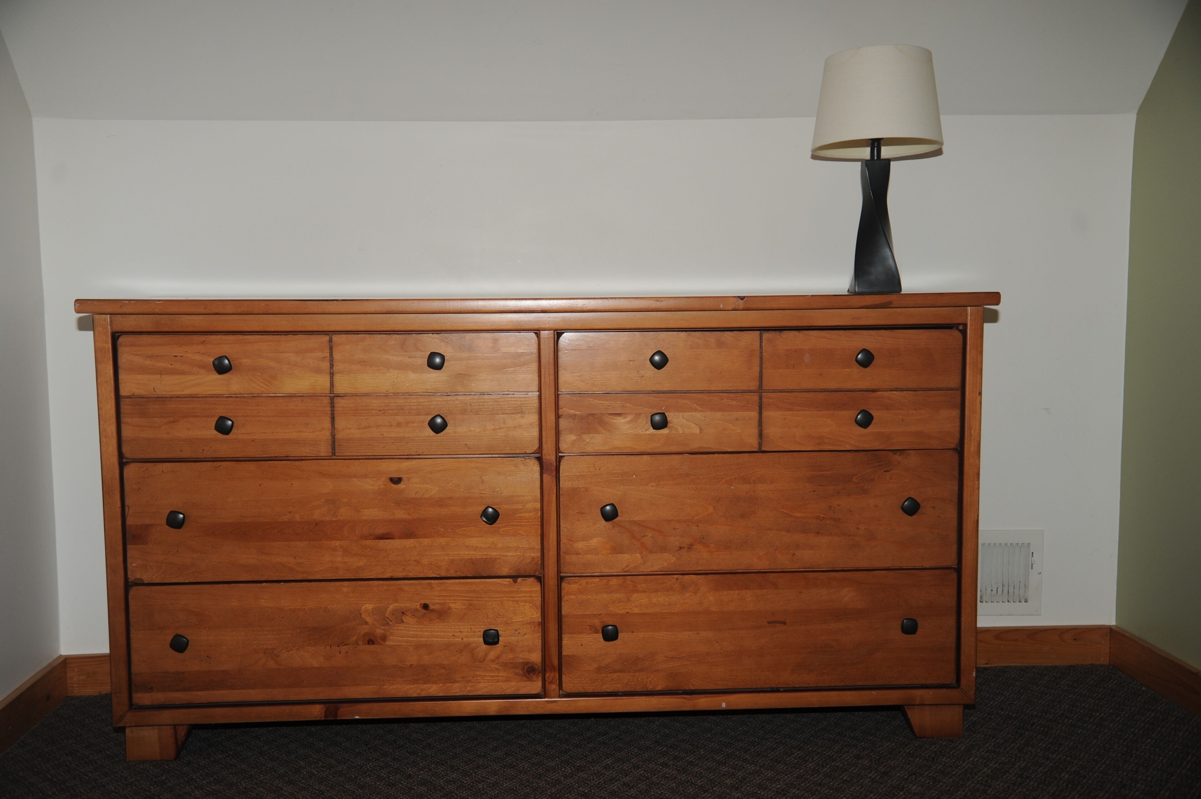 A wooden dresser with multiple drawers and a lamp on top, positioned against a plain wall with a vent below.