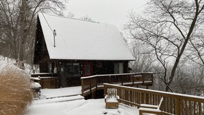 Una cabaña de madera con un porche cubierto de nieve y una puerta roja, rodeada de árboles.