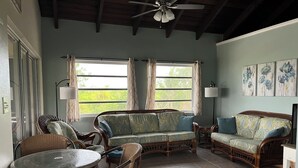 A cozy living room with wicker furniture, a round glass table, and two windows with curtains.
