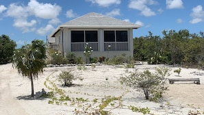 A two-story house with a large window and a balcony, surrounded by a sandy landscape and sparse vegetation.