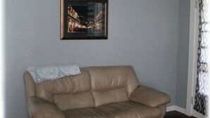 A beige leather sofa with a patterned throw pillow against a light gray wall. A framed picture of a cityscape at night hangs above the sofa. - Beautiful New Orleans Gem Close To Downtown (New Orleans)