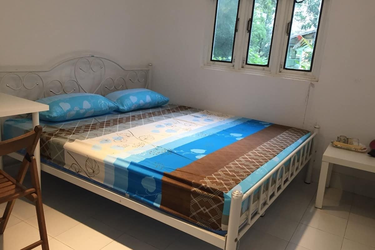 A single bed with a patterned bedspread, a wooden chair, a small table, and a window with a view of greenery.