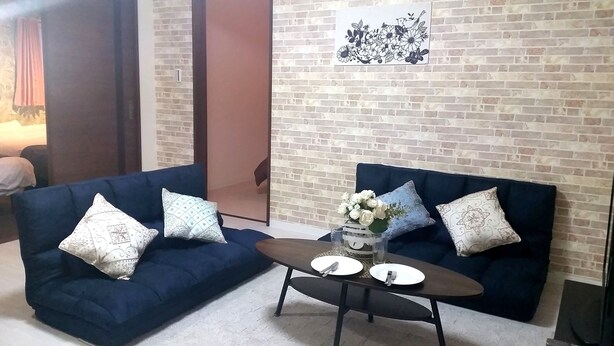 A room with two dark blue sofas with patterned cushions, a small round table with a vase of white flowers, and a brick wall with a floral artwork. - Mansion Kawata (Osaka)