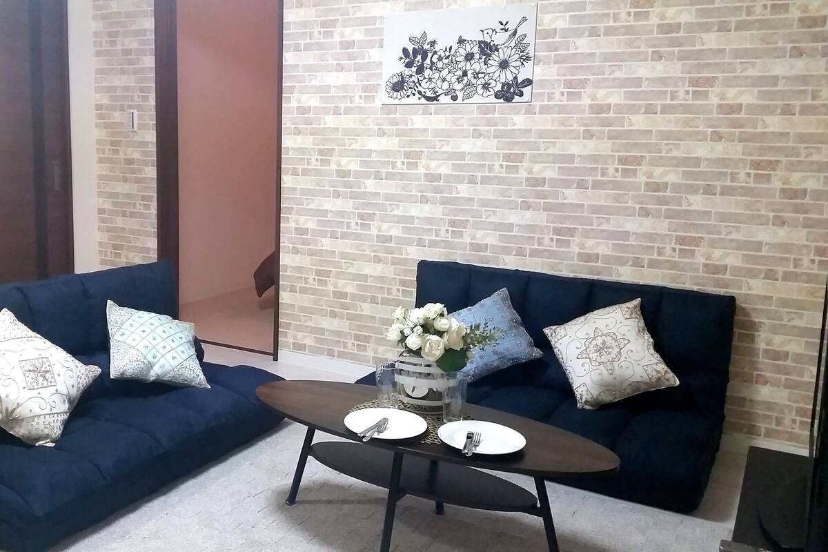 A room with two dark blue sofas with patterned cushions, a small round table with a vase of white flowers, and a brick wall with a floral artwork.