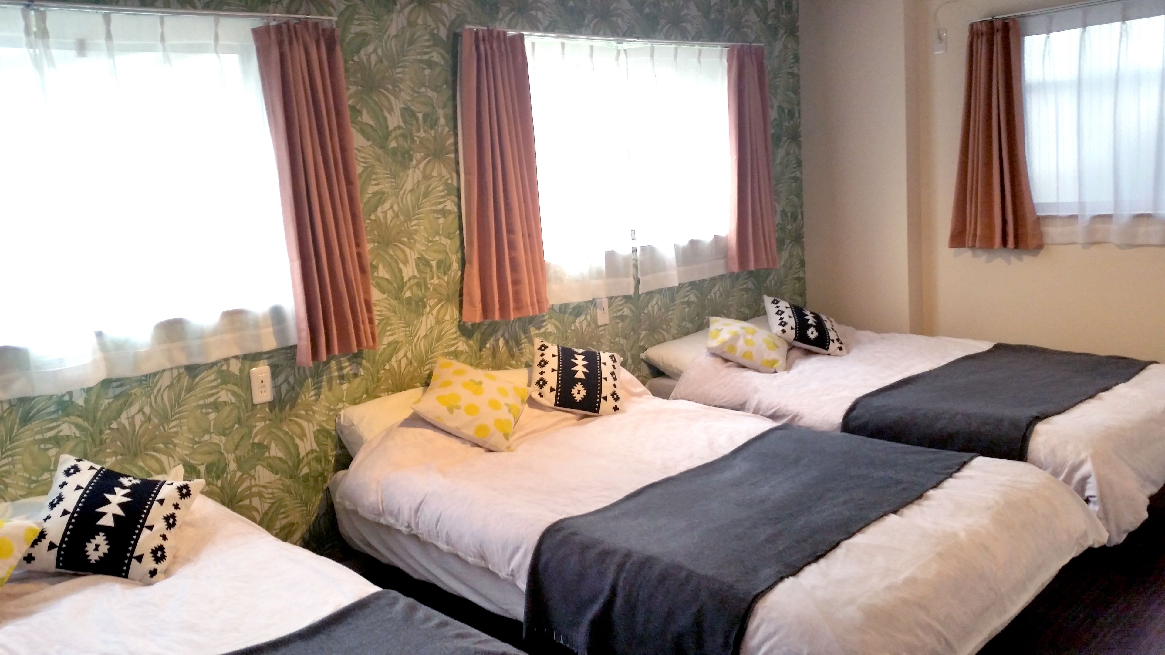 Une chambre d’hôtel avec trois lits, chacun orné d’un oreiller au motif différent. La chambre est décorée d’un papier peint aux motifs tropicaux et de rideaux marron.