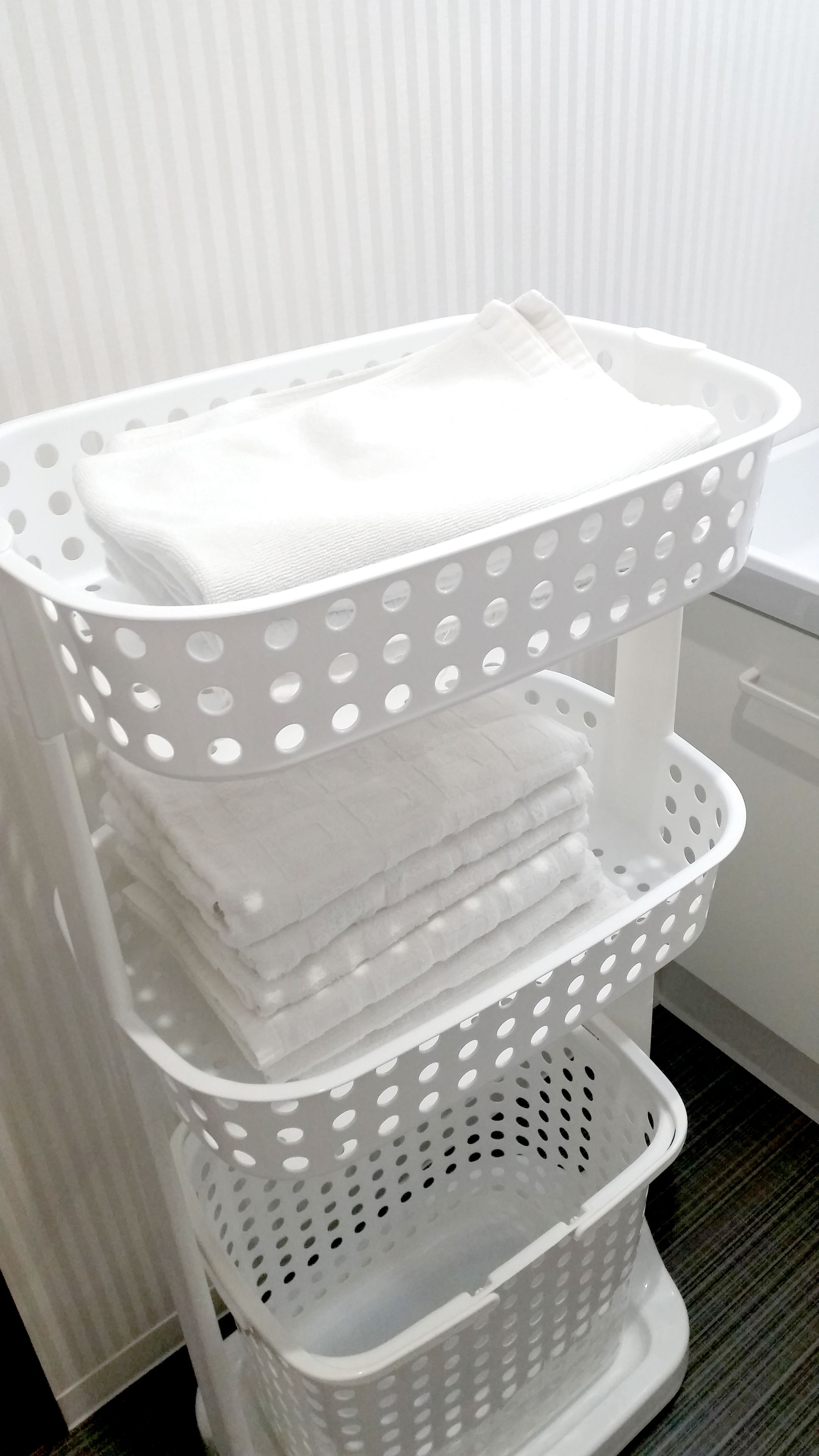 Een witte, drielaagse wasmand met geperforeerde zijkanten, gevuld met opgevouwen witte handdoeken.