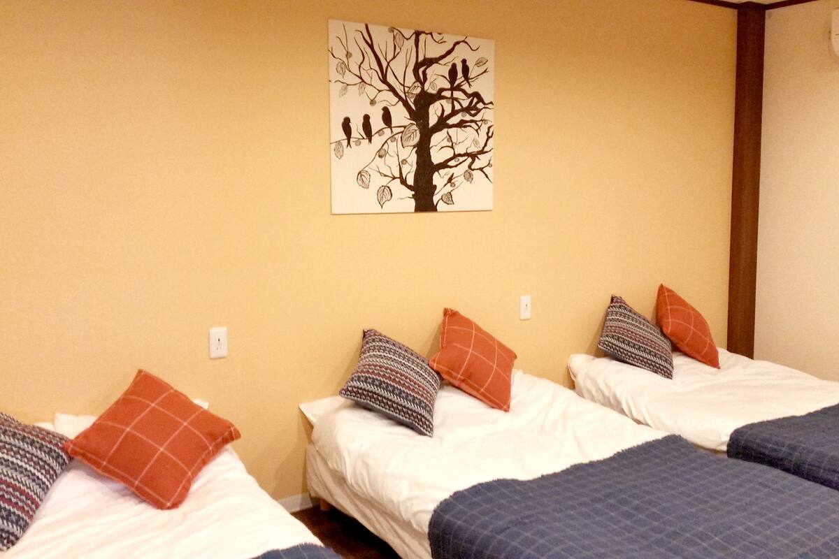 A hotel room with three beds, each with a different patterned pillow and a blue blanket. There is a wall-mounted air conditioning unit and a painting of a tree with birds.
