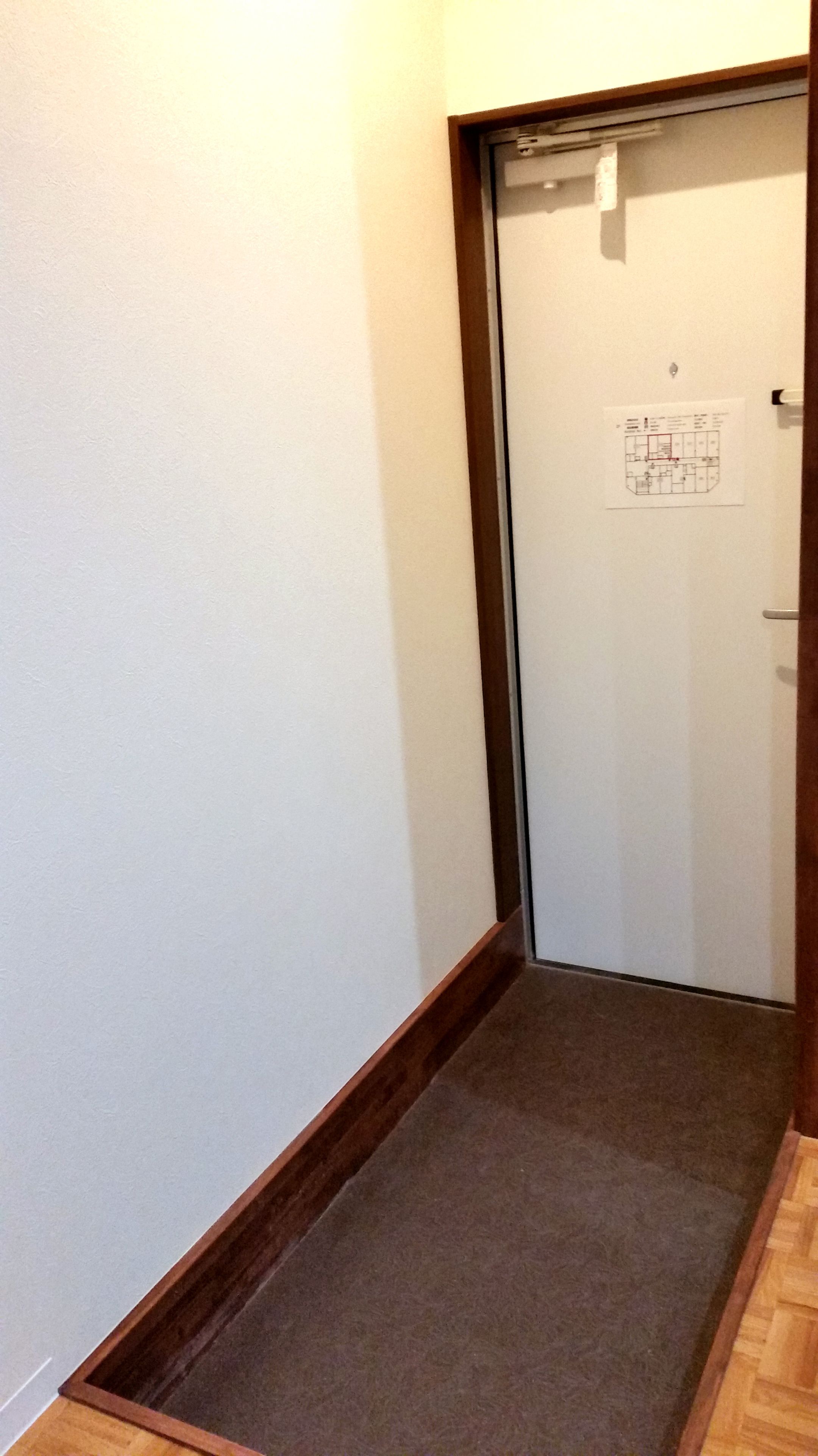 Ein schmaler Flur mit Teppichboden und Holzfußleiste. Eine Tür mit weißem Rahmen ist sichtbar, darüber angebracht eine kleine Schilder mit einem Grundriss.