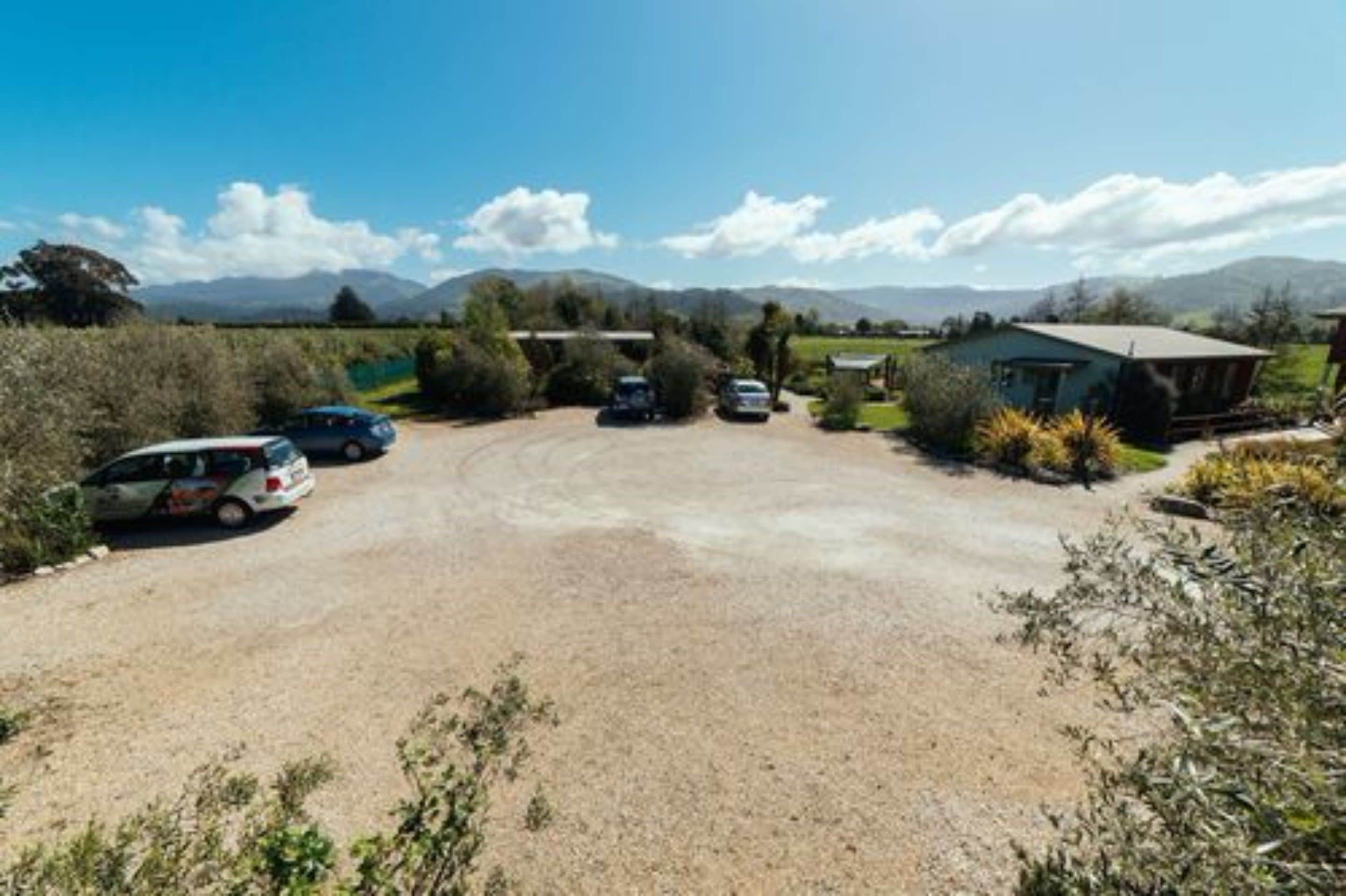 Un estacionamiento de grava con varios coches, un pequeño edificio y un paisaje montañoso al fondo.