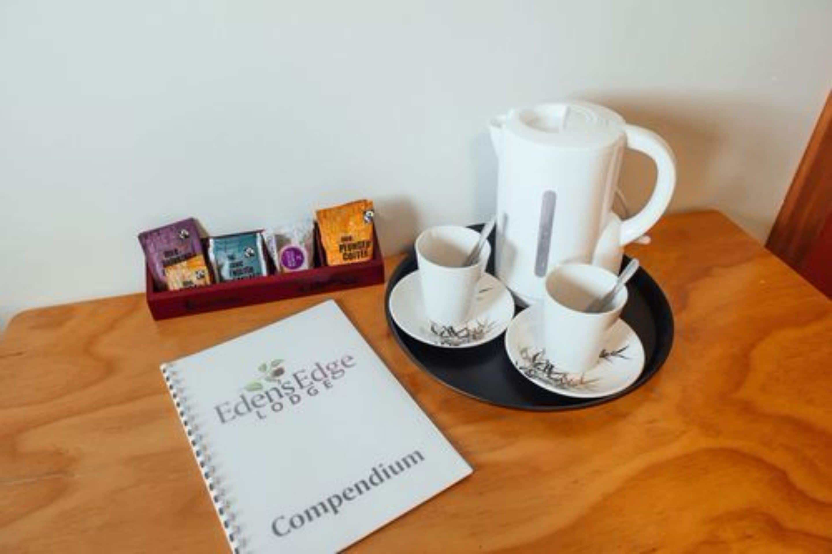A wooden table with a white kettle, two white cups with saucers, and a tray with coffee pods. A notebook titled "Eden's Edge Compendium" is also visible.