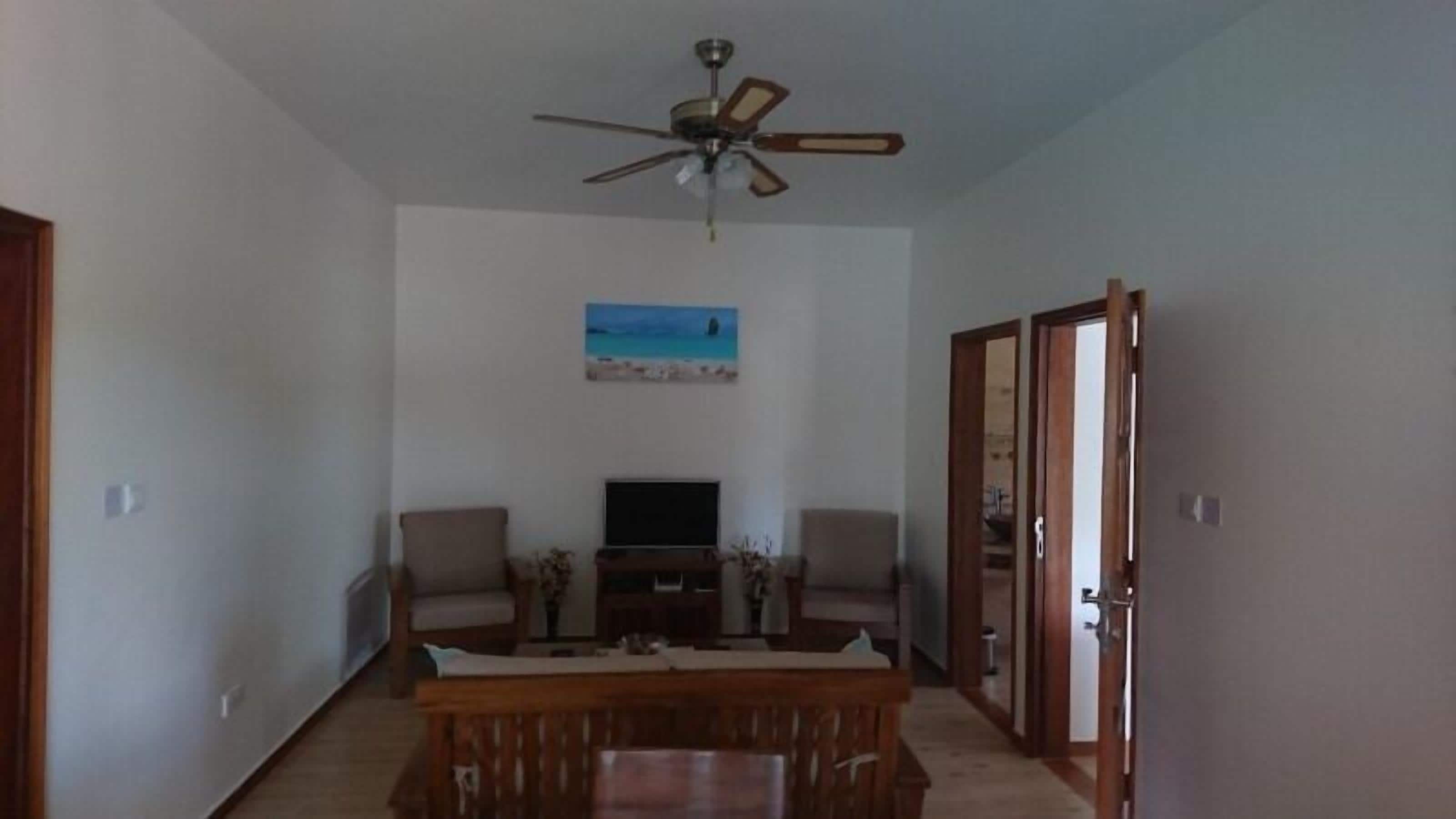 A living room with a ceiling fan, a flat-screen TV, two armchairs, a wooden coffee table, and a painting on the wall.