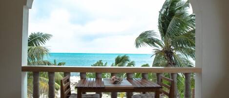 A balcony with a wooden table and chairs, overlooking a beach with palm trees and the ocean.