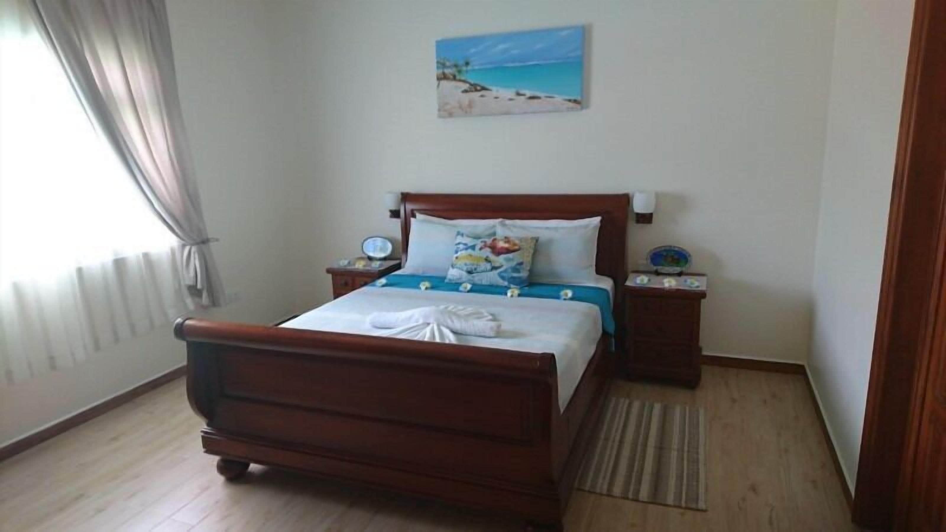 Um quarto com uma cama de madeira, uma mesinha de cabeceira com um relógio, uma mesa de cabeceira com uma luminária e um quadro com tema de praia na parede.