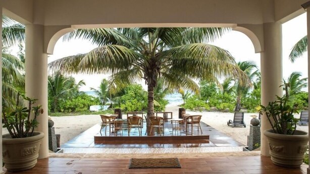 A covered outdoor seating area with a pool, palm trees, and a view of the beach.