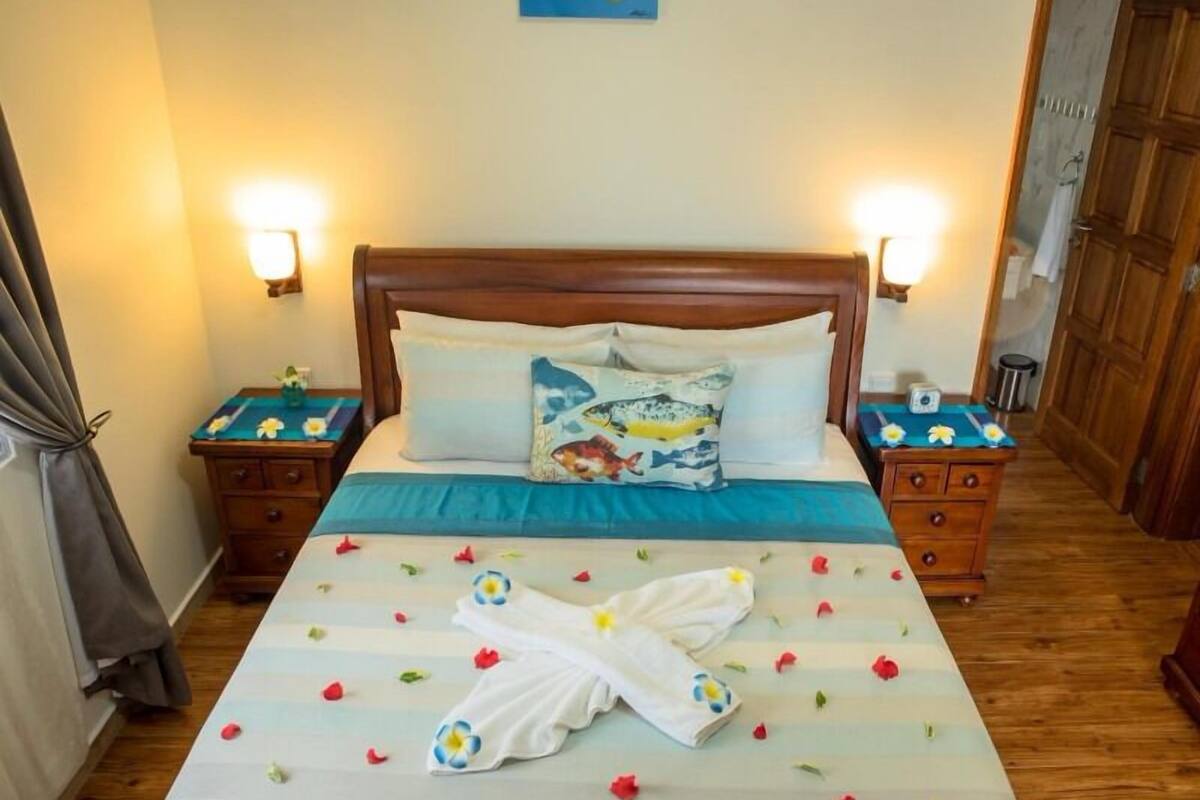Un lit bien fait, avec une couvre-lit bleue et des oreillers blancs, dont un oreiller décoratif orné d’un motif de poisson. On y trouve deux tables de chevet avec des lampes, une armoire en bois et un rideau sur le côté gauche.