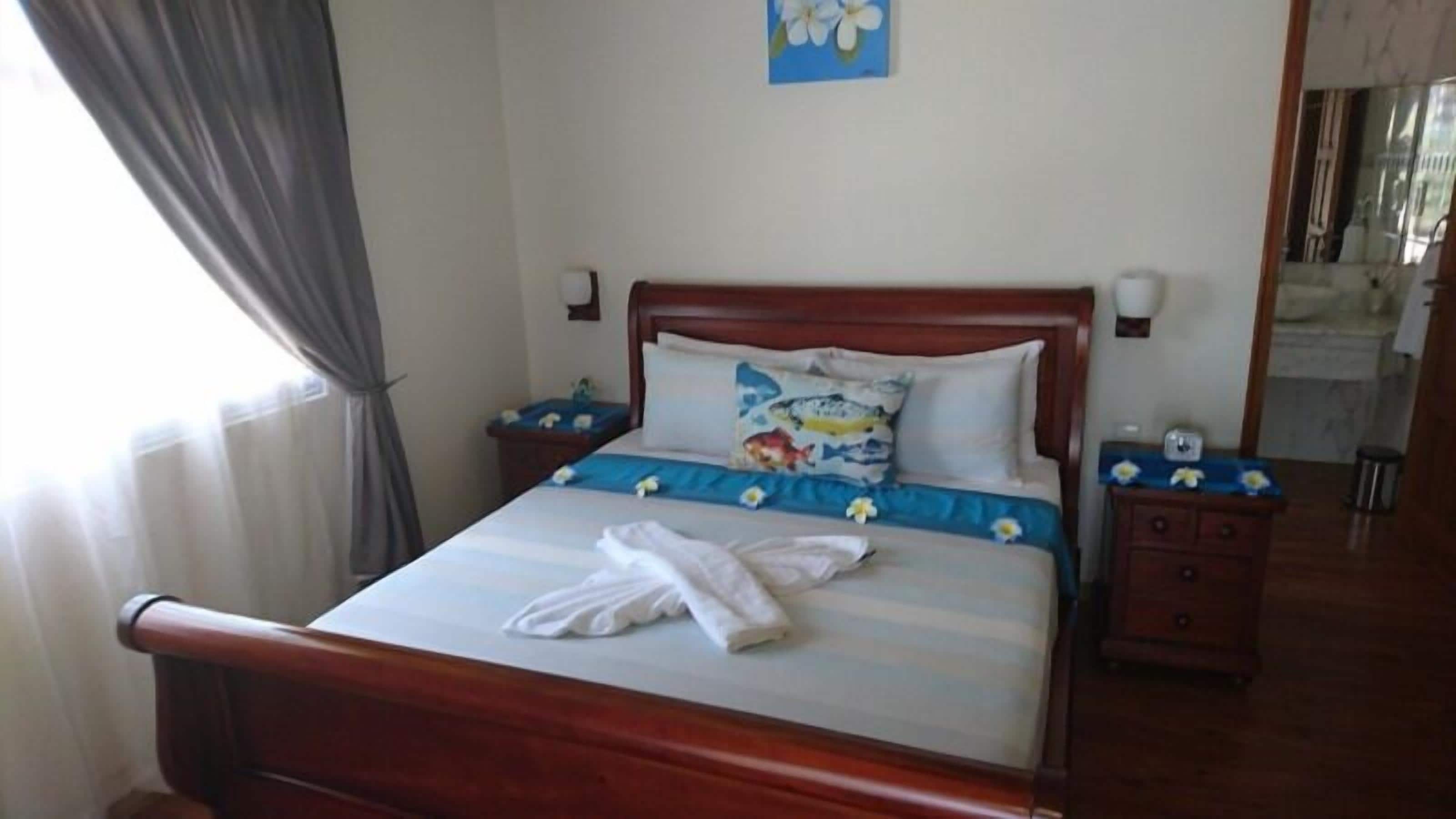Una cama bien hecha con colcha azul y blanca a rayas, una toalla blanca y una almohada estampada con motivos florales. Hay dos mesitas de noche con lámparas, un cubo de basura pequeño y una lámpara de pared.