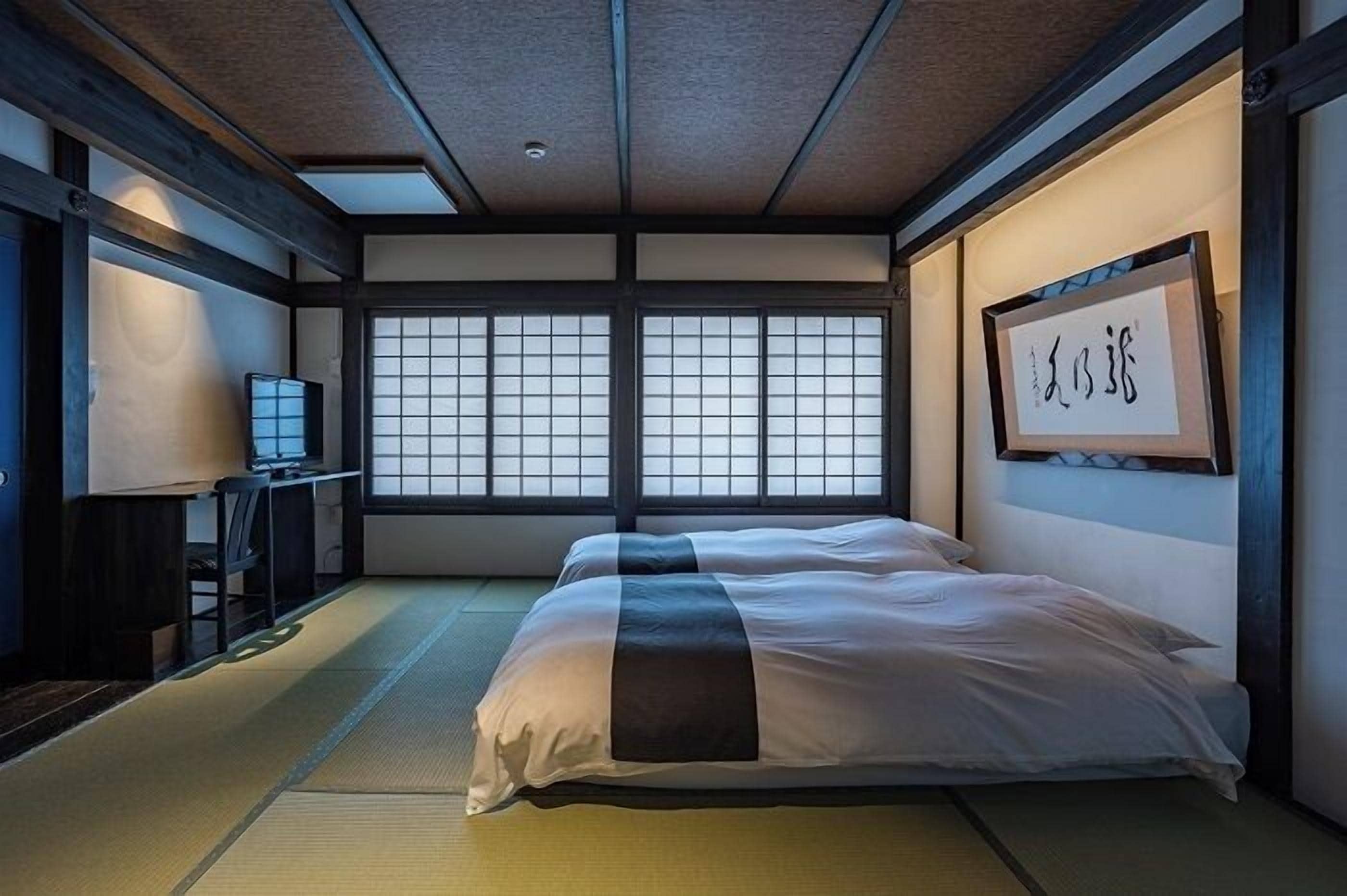A traditional Japanese room with tatami flooring, a bed with white bedding, a television, and a framed calligraphy piece on the wall.