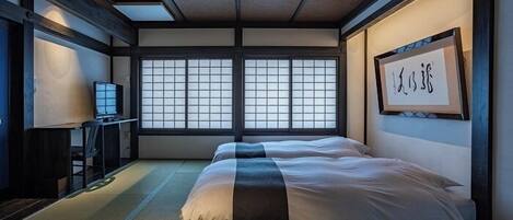 A traditional Japanese room with tatami flooring, a bed with white bedding, a television, and a framed calligraphy piece on the wall.