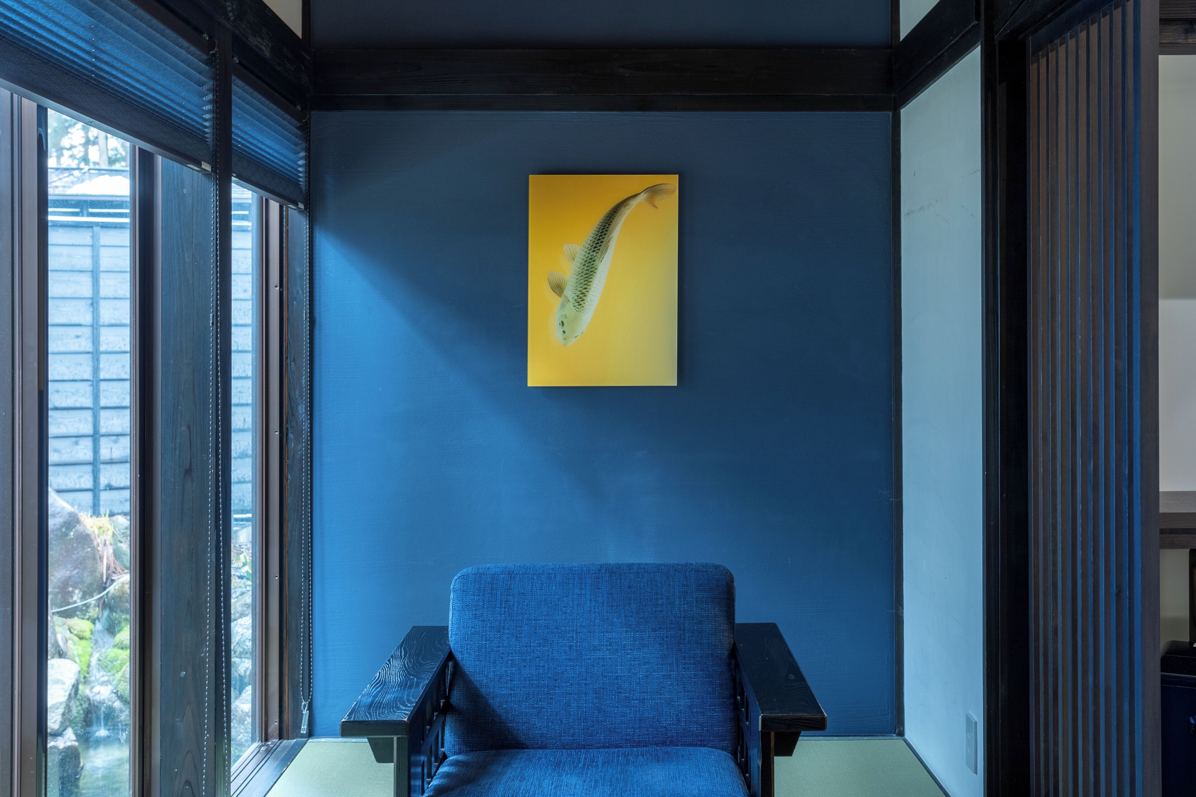 A room with a blue chair, a yellow painting of a fish, and a view through large windows.