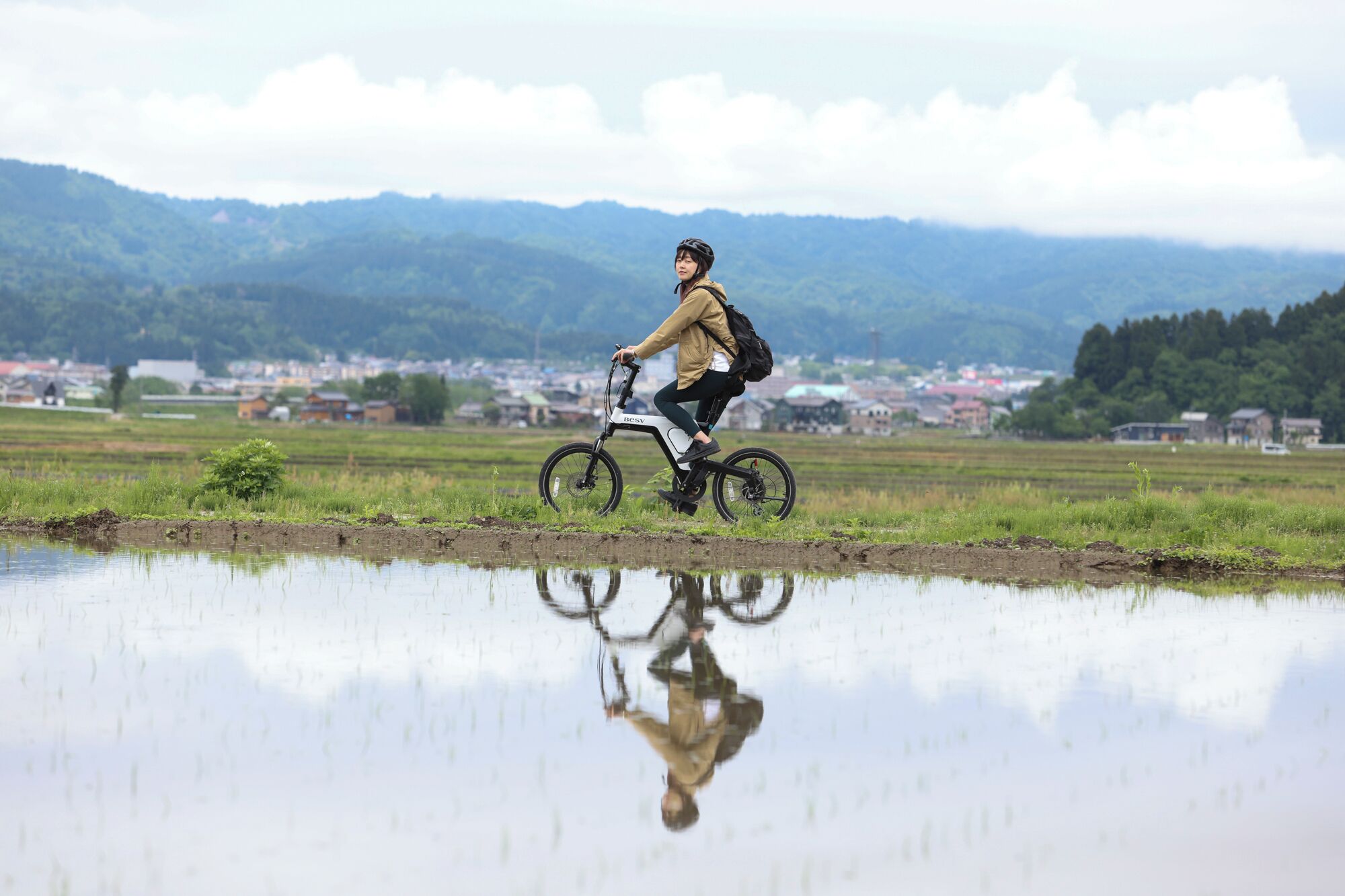 Una persona pedaleando una bicicleta por un campo inundado, con un pueblo al fondo.