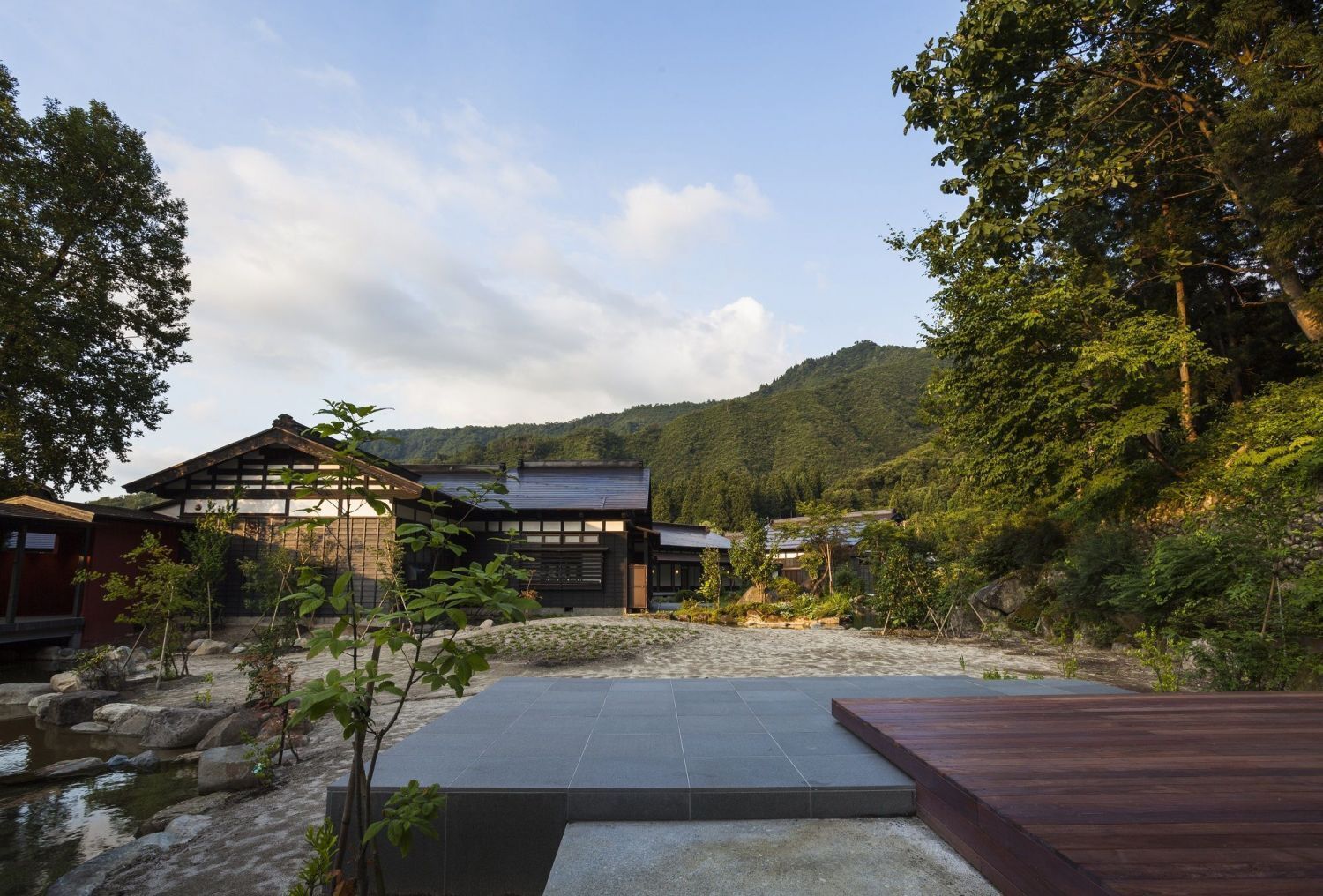 Uma casa japonesa tradicional com um pátio de pedra, rodeada por árvores e colinas.