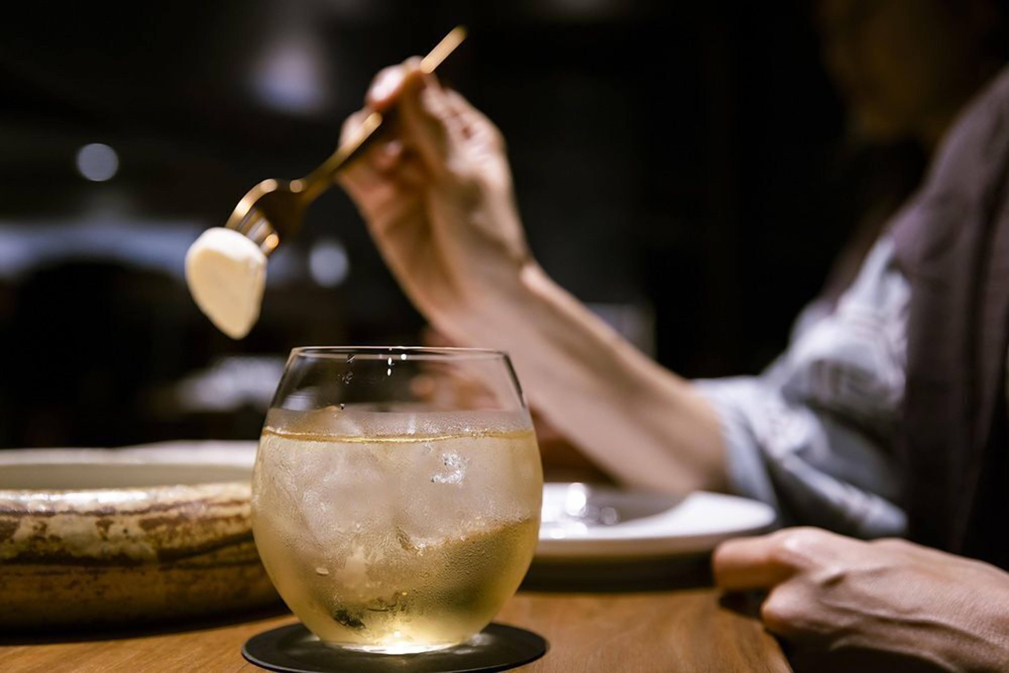 Un verre de vin blanc avec des glaçons, posé sur un dessous de verre, une cuillère, et la main d’une personne tenant la cuillère.