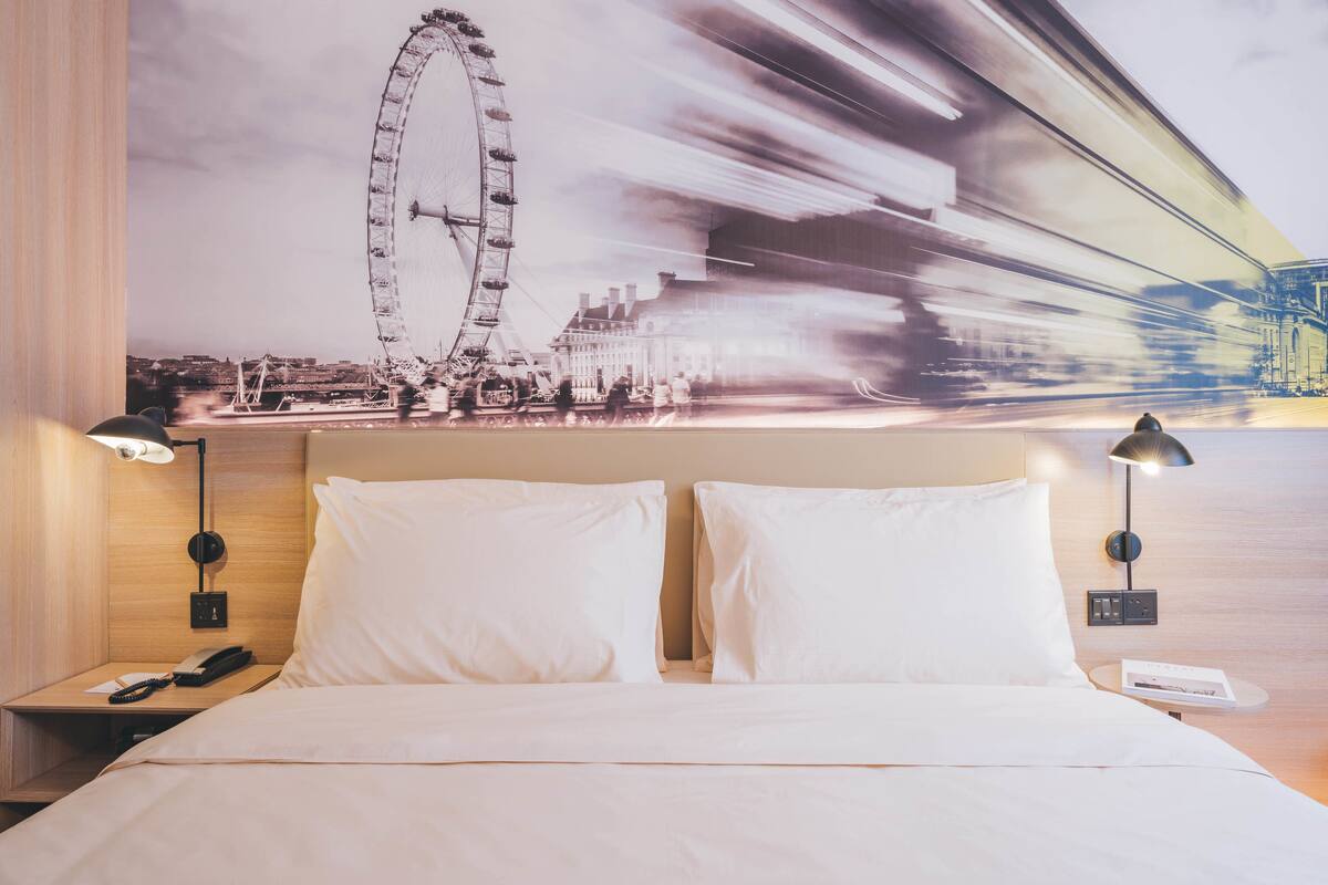 A hotel room with a bed, two bedside tables, a nightstand, a lamp, and a wall-mounted phone.