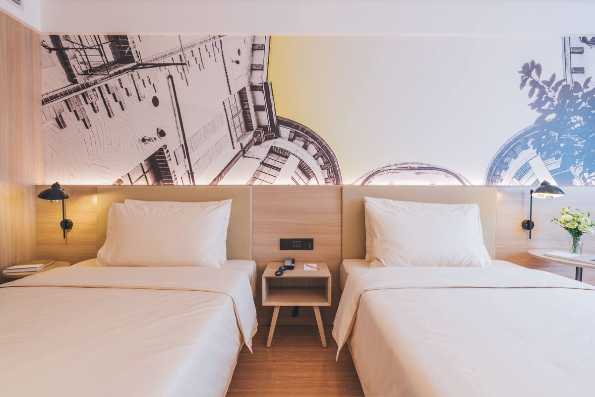 A hotel room with two beds, a nightstand, a wall-mounted control panel, and a small table with a vase of flowers.